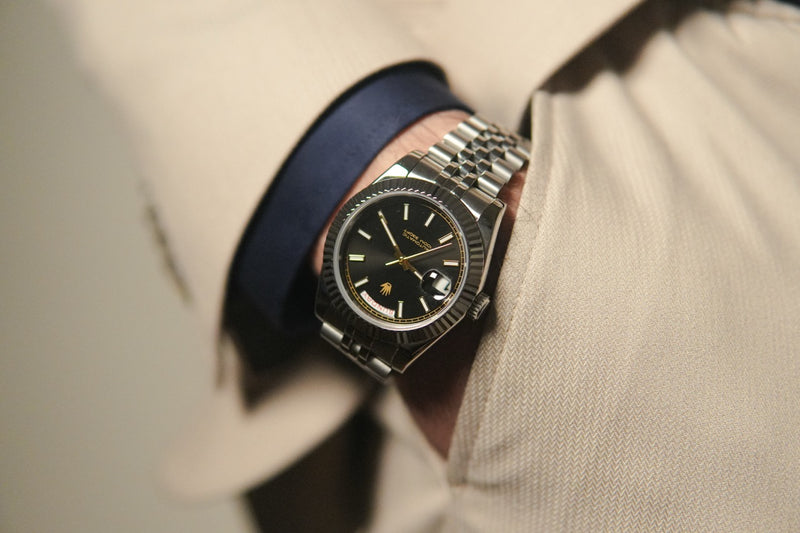 Valenne left-handed automatic watch worn on the right wrist with a tan suit, highlighting the black sunburst dial.