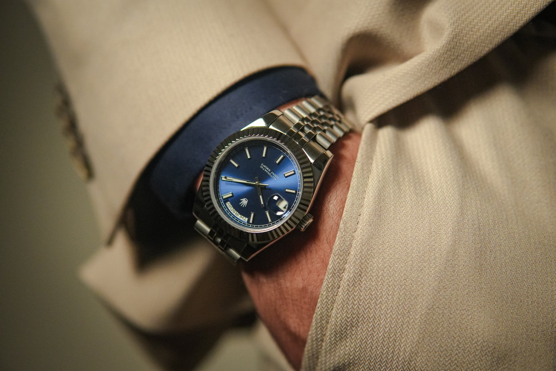 Close-up of a person wearing a watch with a blue face and metallic strap. Life style shot with beige suit